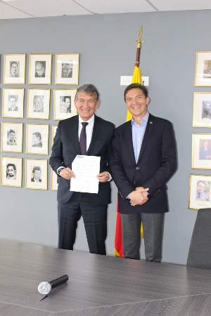 Dos ejecutivos sonrientes de pie en una oficina, sosteniendo un documento firmado, con una bandera y retratos enmarcados detrás.
