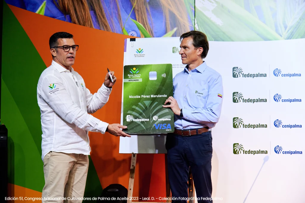 Dos hombres, uno de ellos con gafas, sostienen una tarjeta de débito VISA promocional gigante de color verde con el logo de Fedepalma.
