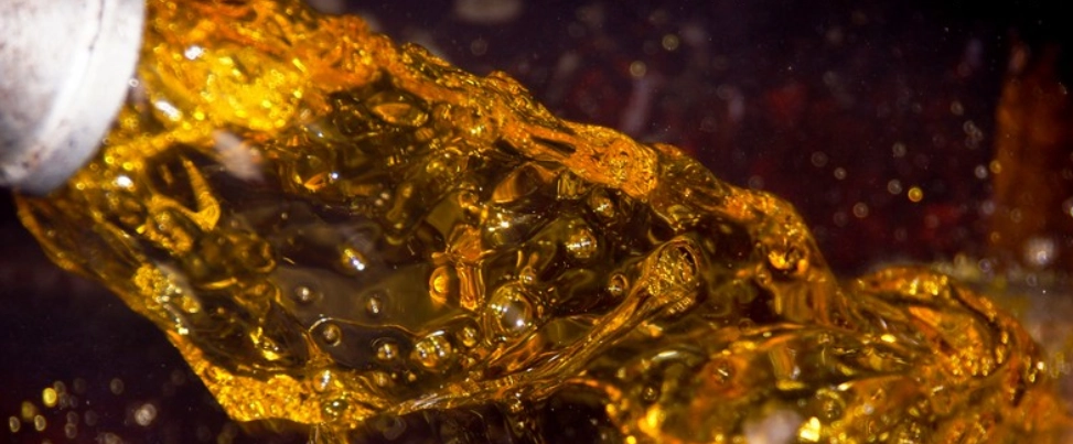 Primer plano de un chorro espeso y burbujeante de aceite de palma caliente, de color naranja/ámbar, destacándose al verterse desde una tubería blanca.