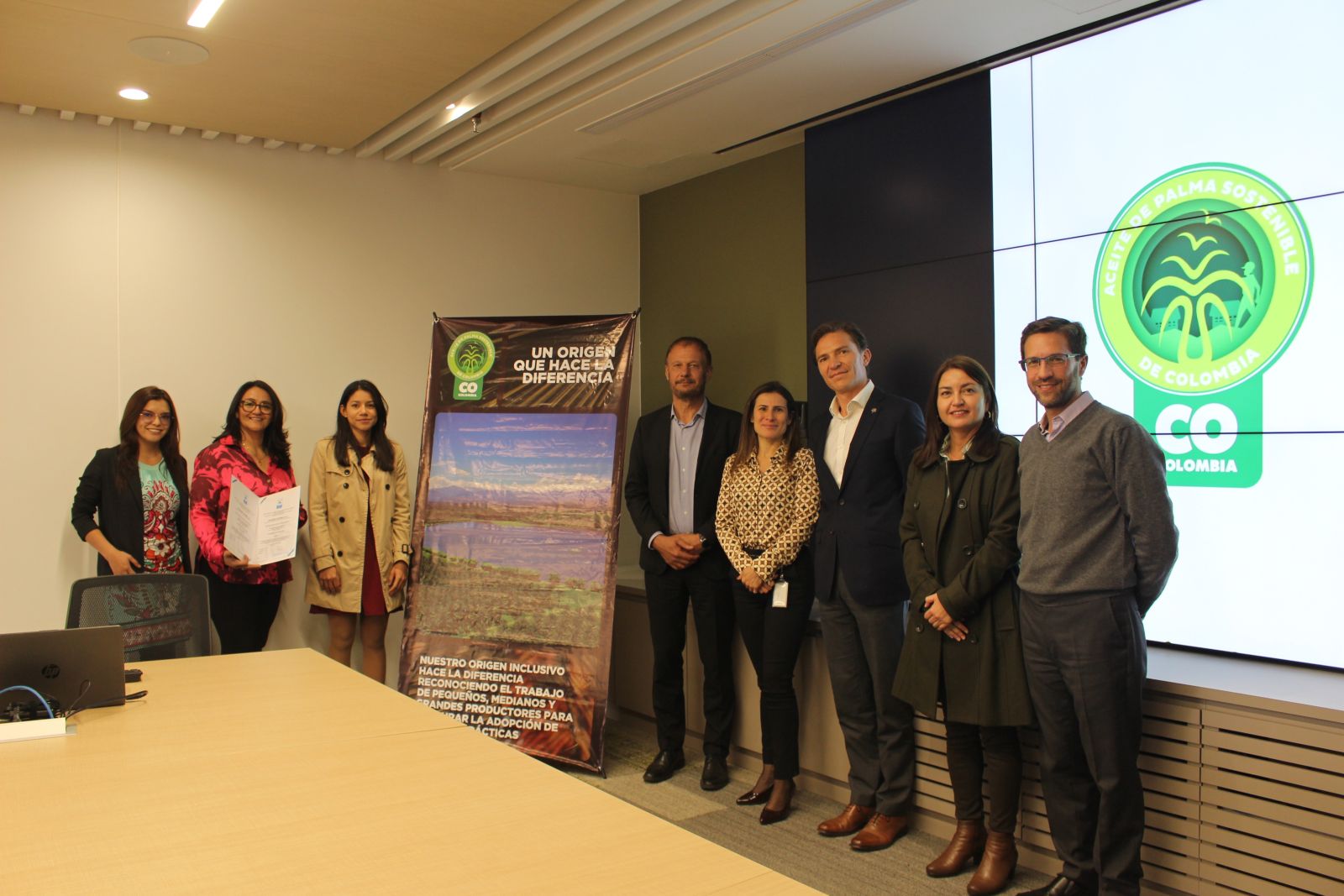 Ocho personas posando en una sala de reuniones frente a un banner y una pantalla con el logo de 'Aceite de Palma Sostenible de Colombia CO'