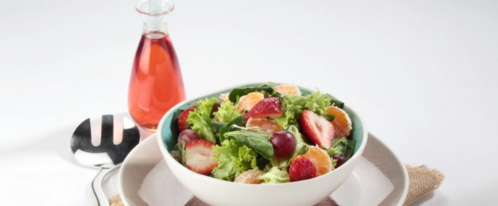 "Presentación de un tazón de ensalada gourmet de frutas y hojas verdes, servido sobre un plato con una textura de arpillera. A un lado, se encuentra una botella de aderezo de color rosa brillante y utensilios de plata