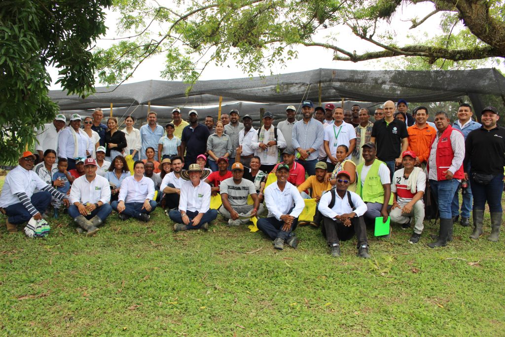 Foto grupal de aproximadamente 50 a 60 personas, incluyendo agricultores y personal de campo, posando al aire libre bajo la sombra de un árbol.