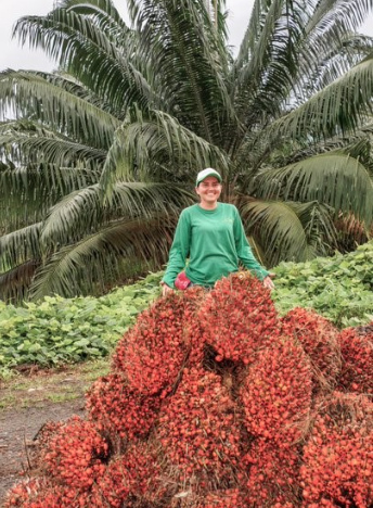 "Mujer sonriendo y vestida de verde de pie detrás de una gran pila de racimos de frutos de palma de aceite cosechados, con una palmera detrás."