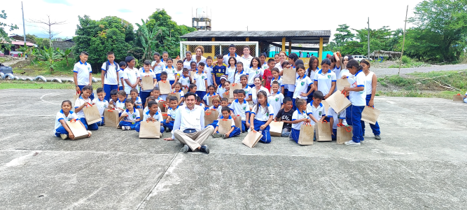 Grupo de escolares en uniforme y adultos posando en una cancha de cemento con bolsas de papel, durante un evento de entrega de útiles o regalos