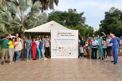 Gran grupo de personas, posando ante el cartel del 'Programa Integral de Salud: Palmas Para Una Sola Salud' al aire libre.