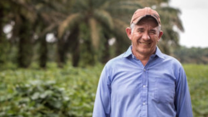 Hombre sonriente de mediana edad, de pie en un campo verde con palmas de aceite desenfocadas en el fondo.