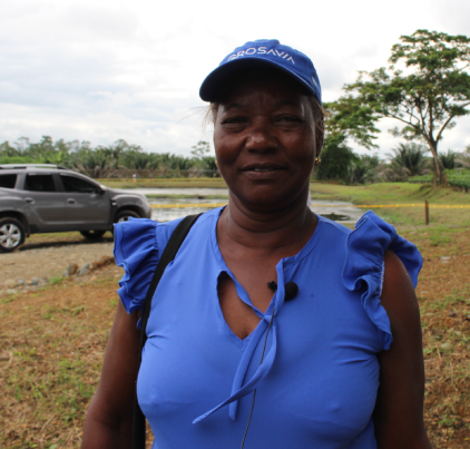 Retrato de una mujer sonriendo, con gorra azul de AGROSAVIA, en un campo de cultivo con un vehículo detrás.