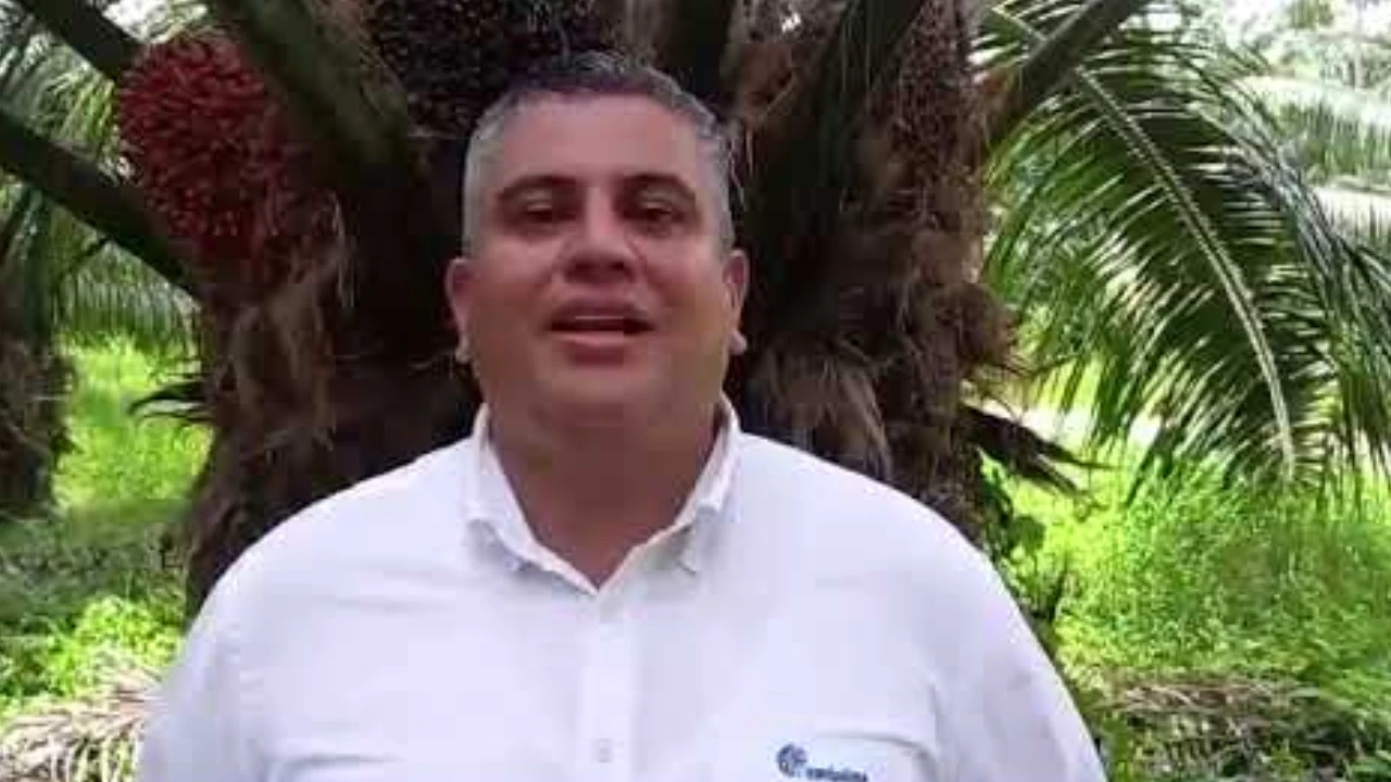 Hombre sonriente, con camisa blanca, frente a un tronco de palma de aceite con frutos