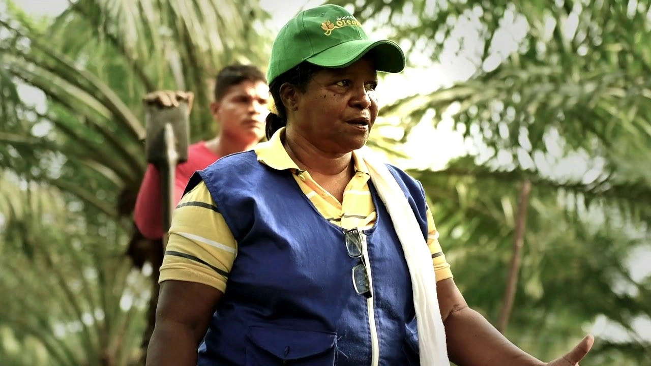 Mujer con gorra verde, chaleco azul y camisa amarilla, hablando en un campo de palmas con otra persona en el fondo.