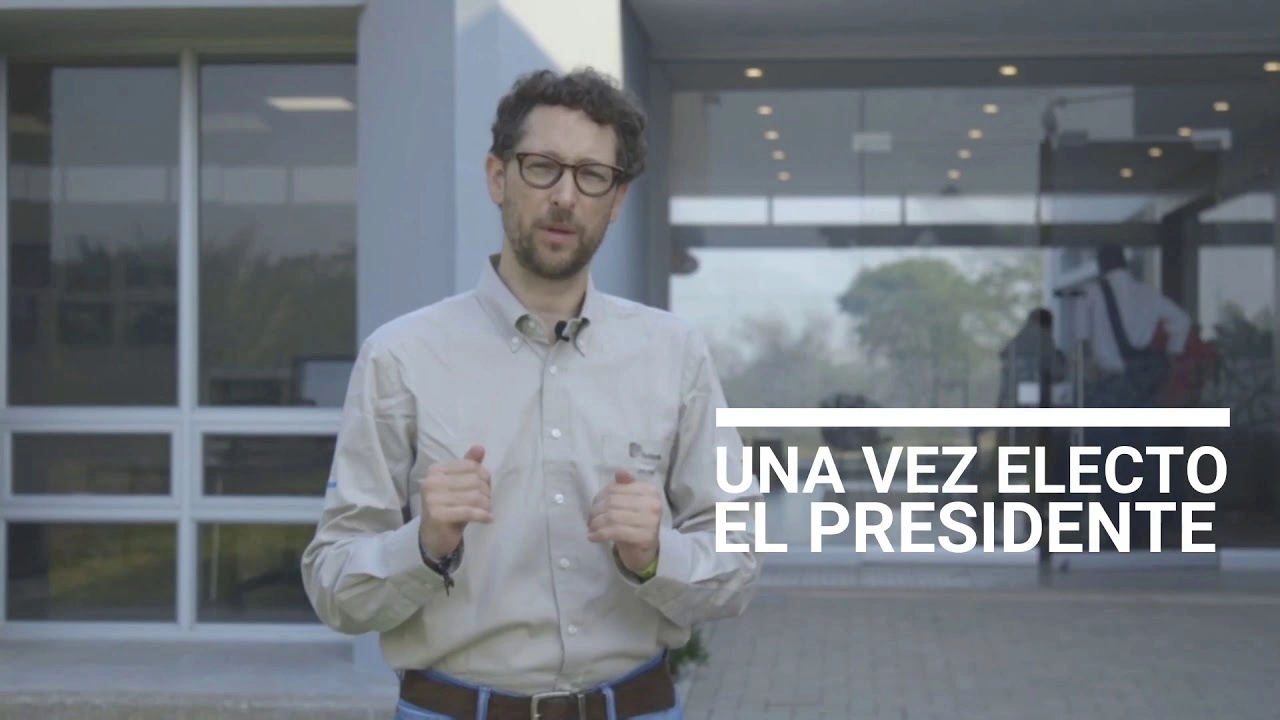 Un hombre con camisa clara y gafas de montura negra de pie frente a un edificio moderno con ventanas de vidrio, con el texto 'UNA VEZ ELECTO EL PRESIDENTE'