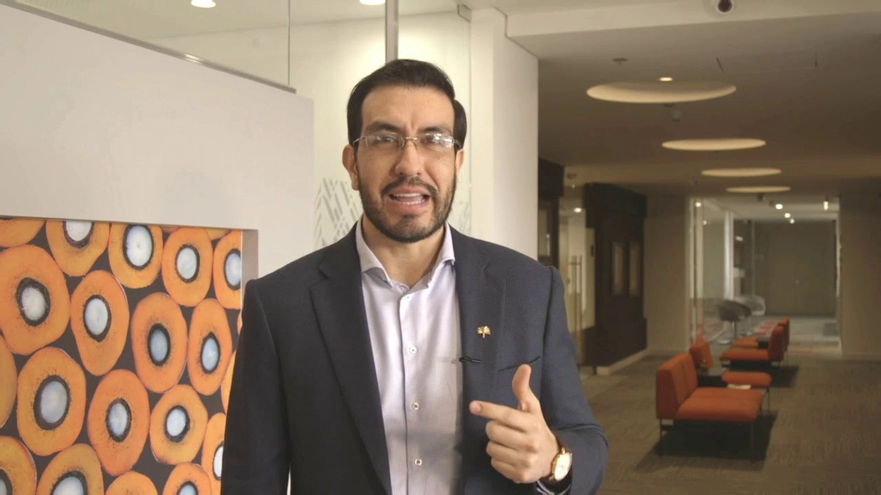 Hombre con barba, hablando en un pasillo de oficina, con un cuadro de frutos de palma.