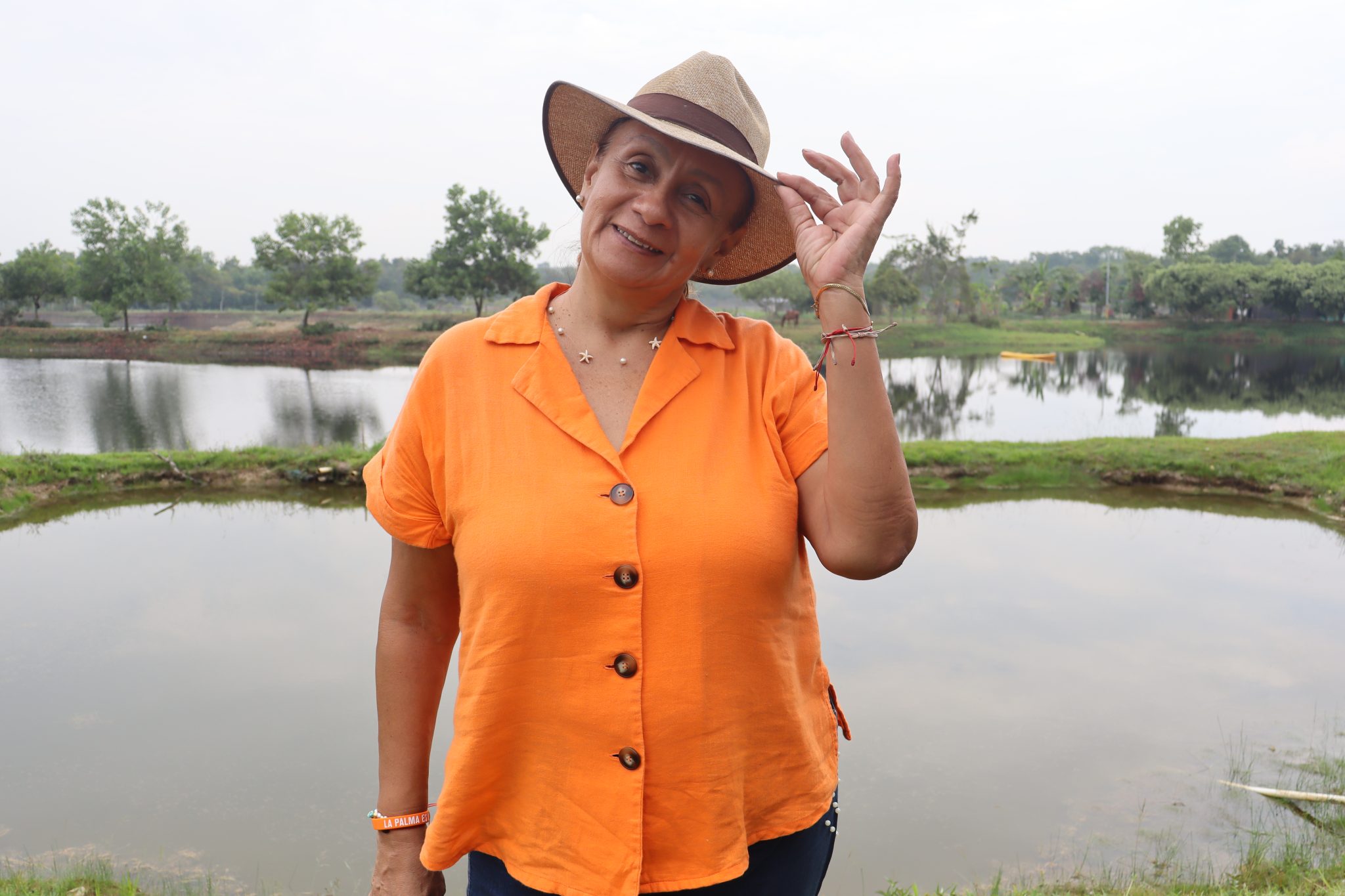Mujer sonriente con camisa naranja y sombrero de paja, posando junto a un lago rodeado de árboles.