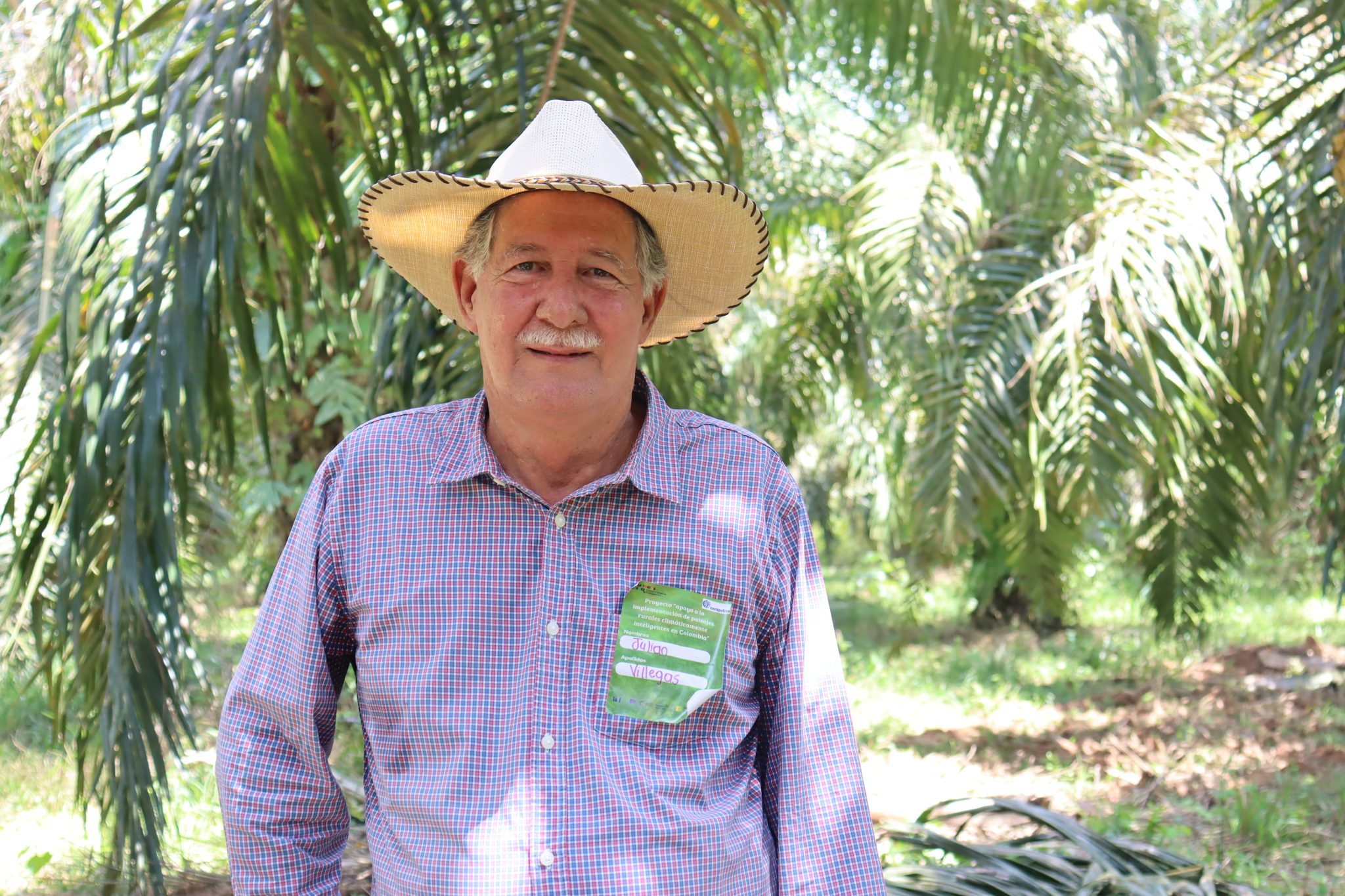 Retrato de un palmicultor sonriendo, con un sombrero y escarapela, en medio de una plantación de palma.