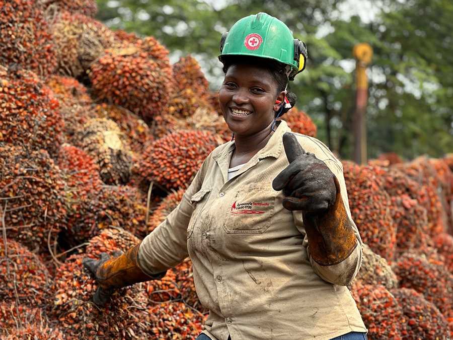 Retrato de una mujer trabajadora sonriente, rente a una gran pila de racimos de frutos de palma de aceite