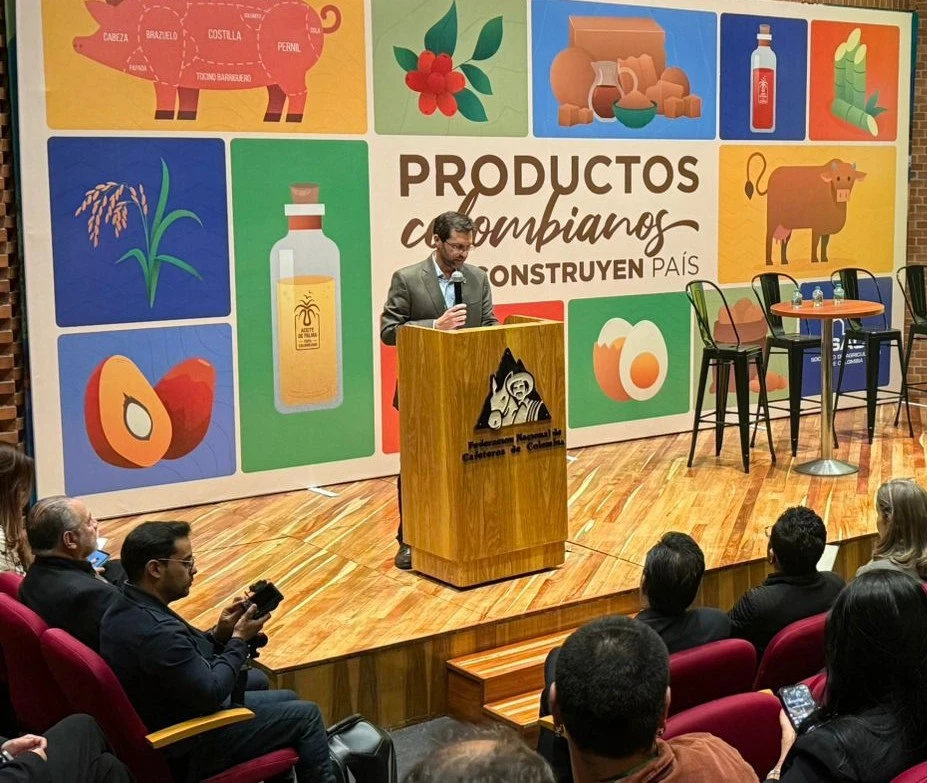 Orador en un podio con el escudo de la Federación Nacional de Cafeteros de Colombia, frente a un mural con ilustraciones de 'Productos Colombianos'