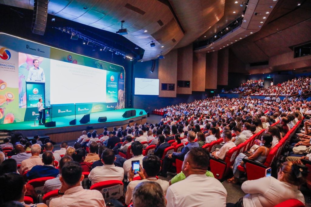 vista panorámica de un gran auditorio lleno de gente, con un orador en el escenario presentando frente a una gran pantalla.