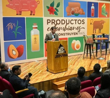 Orador en un podio con el escudo de la Federación Nacional de Cafeteros de Colombia, frente a un mural con ilustraciones de 'Productos Colombianos'