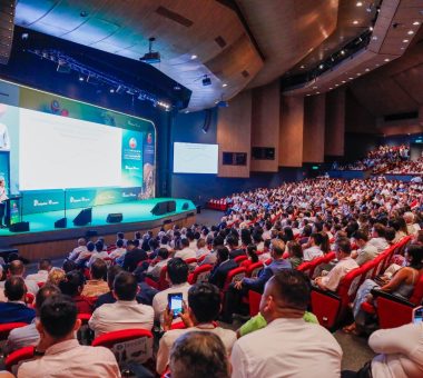 vista panorámica de un gran auditorio lleno de gente, con un orador en el escenario presentando frente a una gran pantalla.