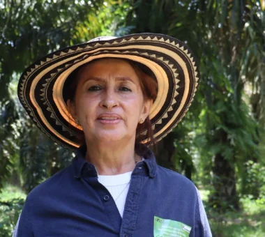 Retrato de una mujer con un sombrero vueltiao tradicional, parada en medio de una plantación de palma de aceite bajo el sol.